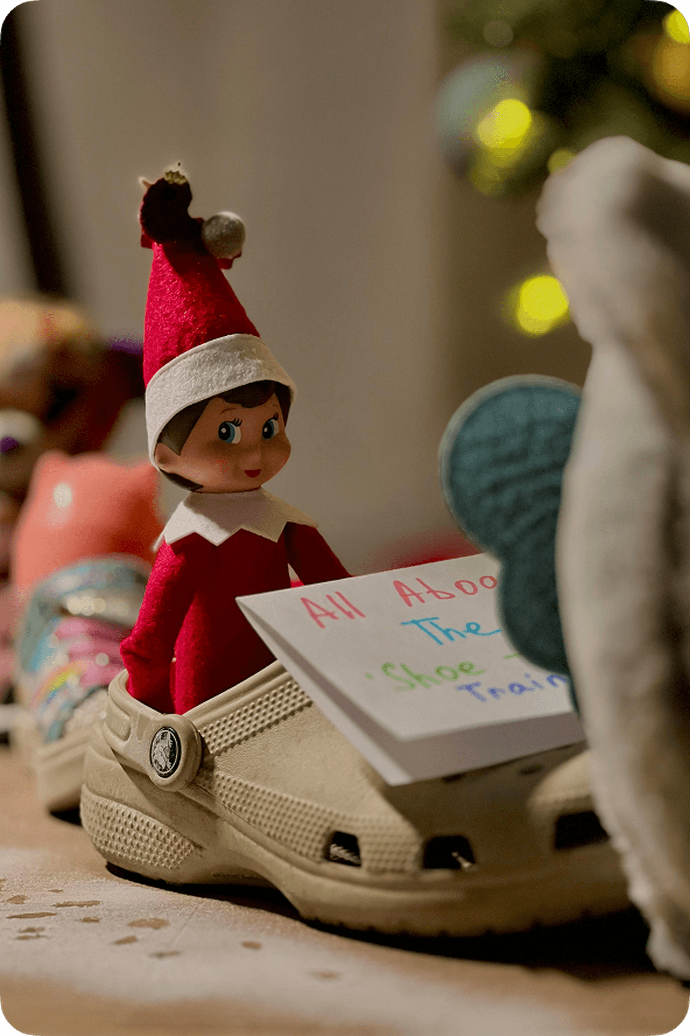 Elf doll in a red outfit sitting on a shoe, holding a book titled "All About The Shoe Train," with blurred Christmas lights in the background.