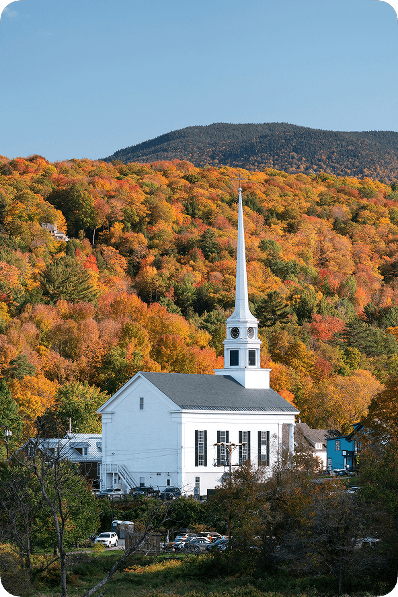 White church with a tall steeple set against vibrant autumn foliage and a mountain backdrop under a clear blue sky.