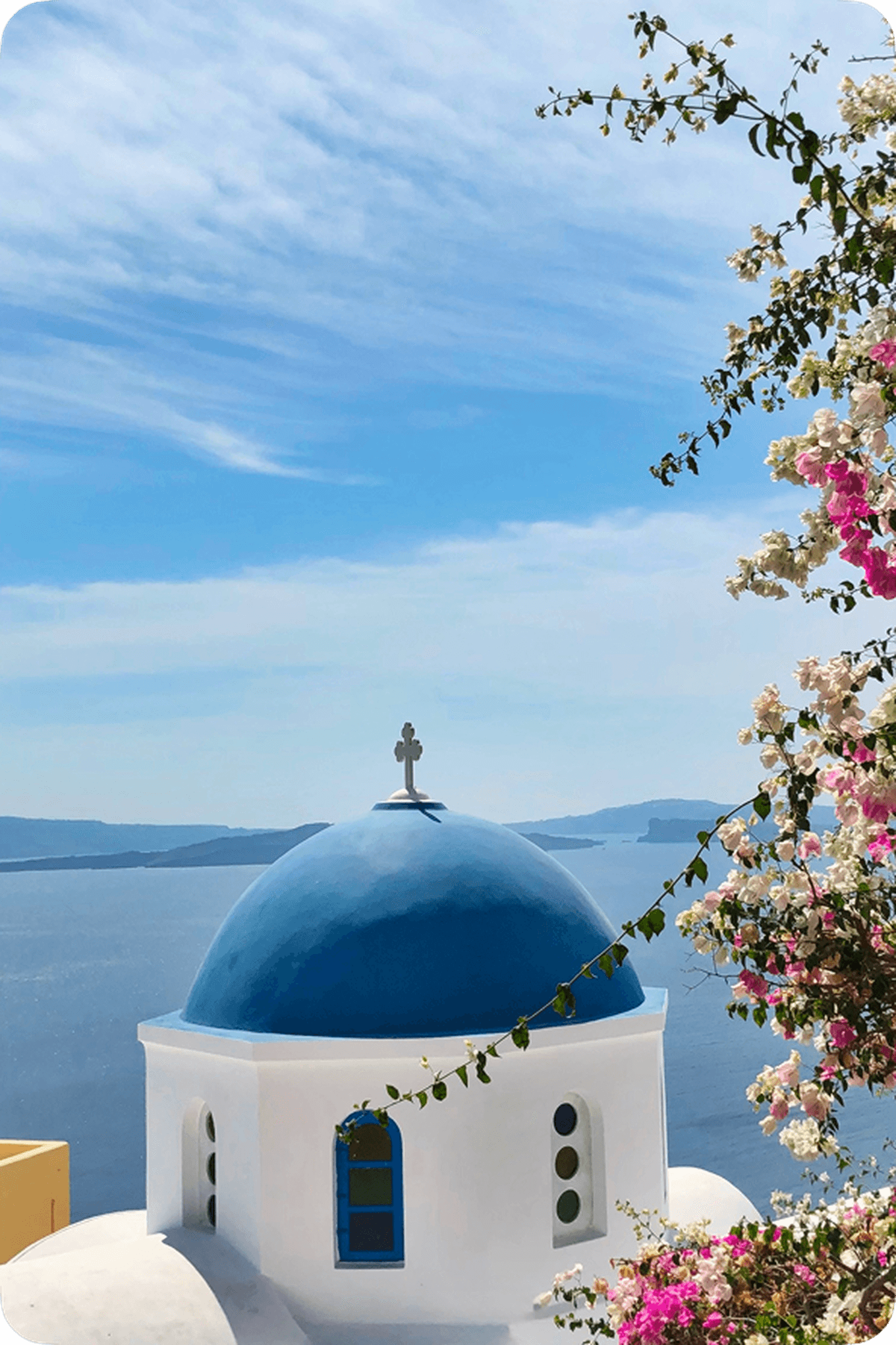 White building with a blue dome and cross overlooking the sea, framed by pink and white flowers, under a clear blue sky.