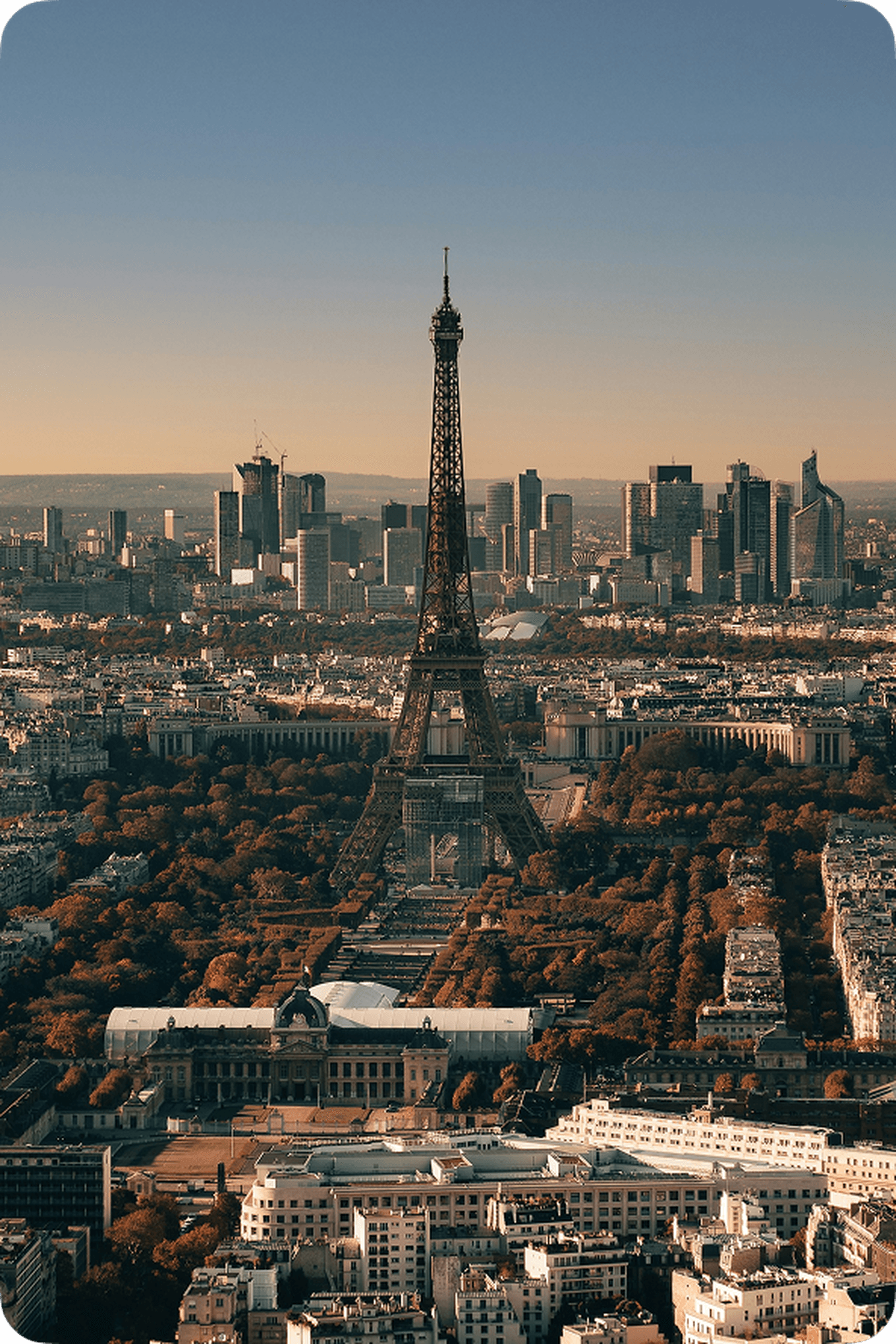Aerial view of the Eiffel Tower surrounded by autumn foliage, with Paris cityscape and modern skyscrapers in the background under a clear sky.