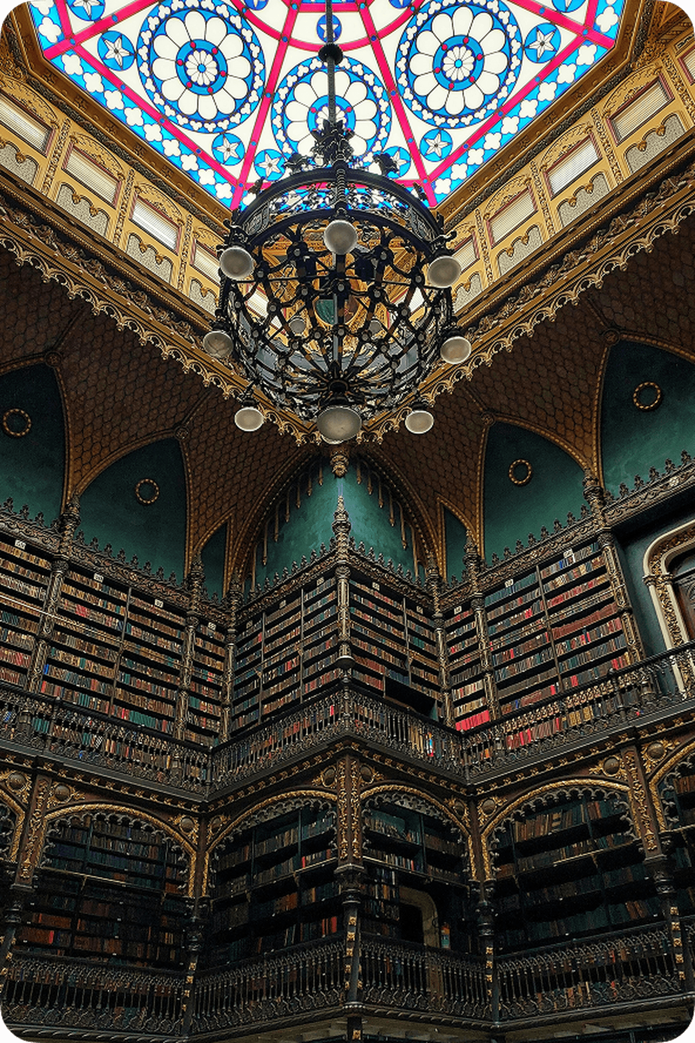 Ornate library interior with tall bookshelves, intricate woodwork, and a large chandelier under a colorful stained glass ceiling.