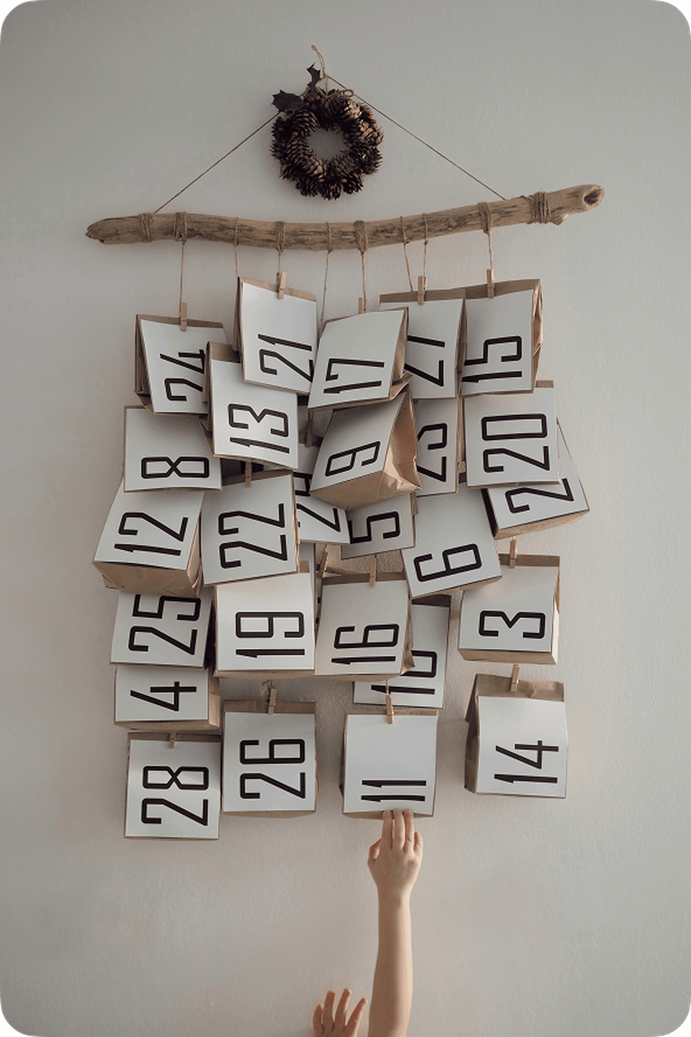 A child reaches for a hanging advent calendar made of paper bags numbered 1 to 28, displayed on a wall with a wooden stick and wreath.