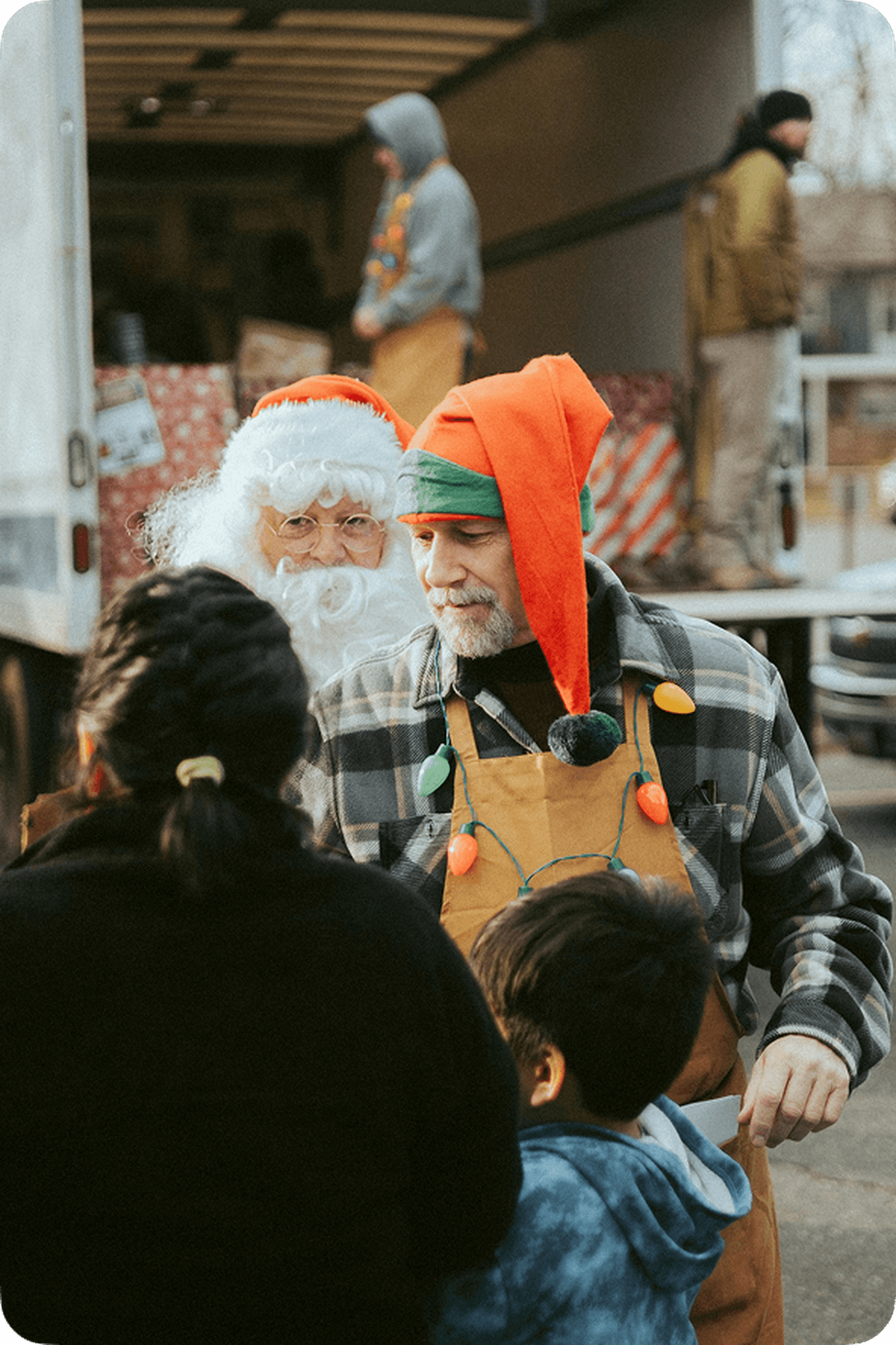 Man in festive hat and apron talks to a woman and child. Santa Claus and others unload a truck in the background.