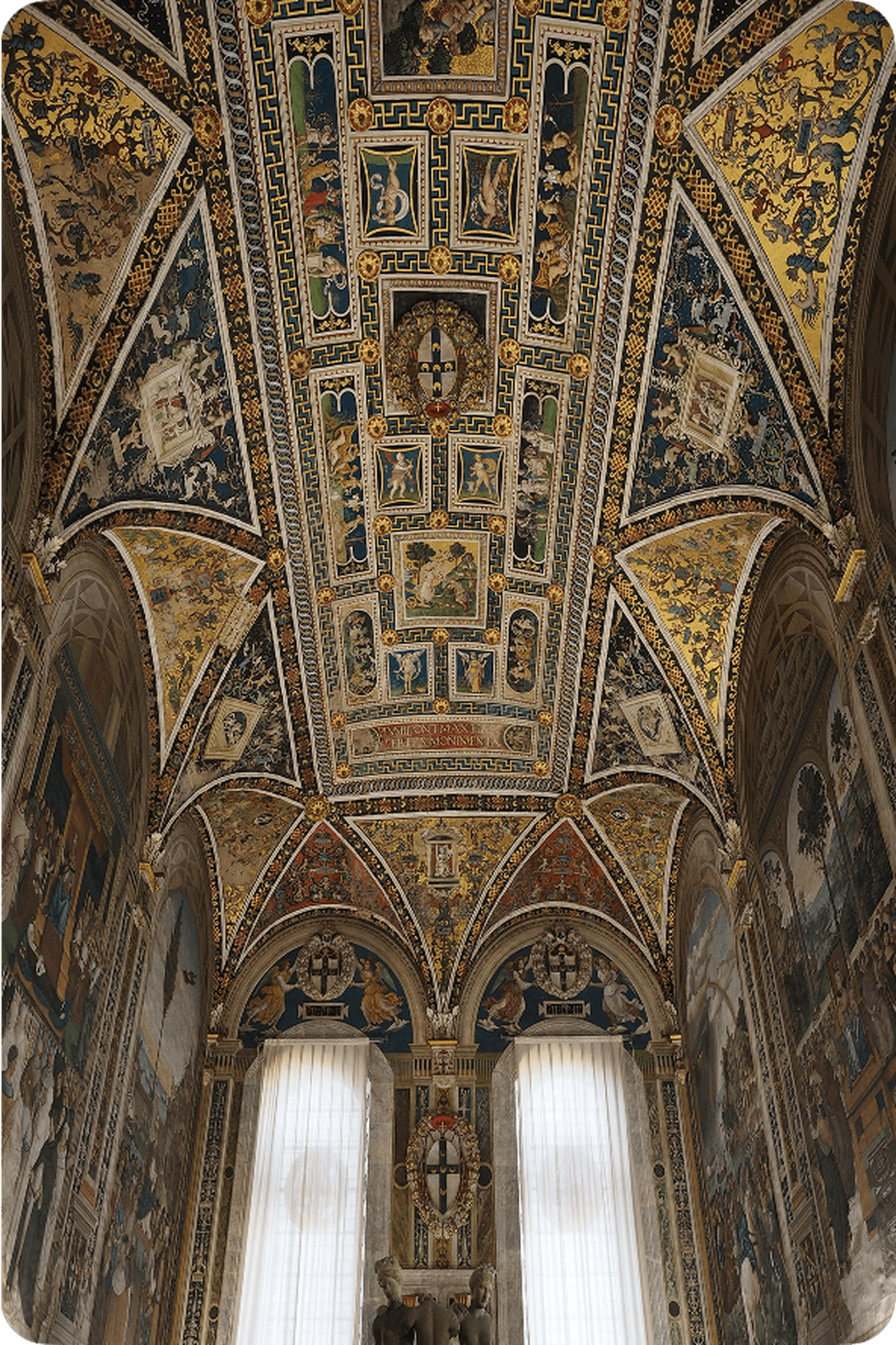 Ornate, vaulted ceiling with intricate frescoes and detailed patterns in a richly decorated room, illuminated by large windows.