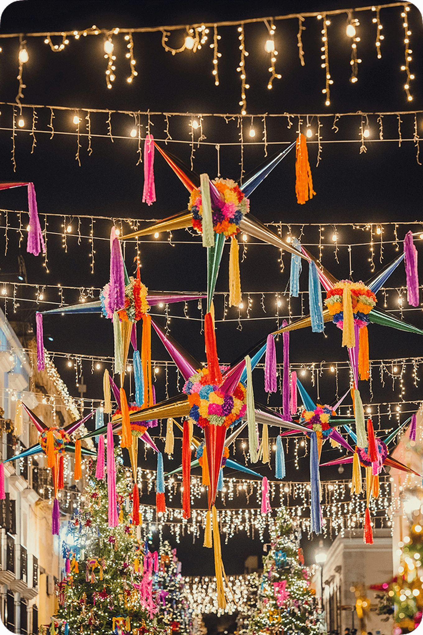 Colorful piñatas and string lights hang above a festive, decorated street at night, creating a vibrant and joyful atmosphere.