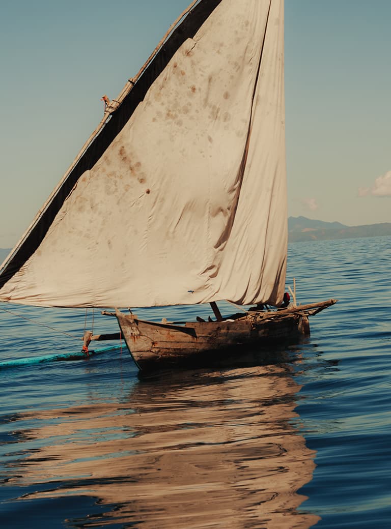 A small wooden sailboat with a large white sail glides over calm blue waters under a clear sky. Distant mountains are visible in the background.