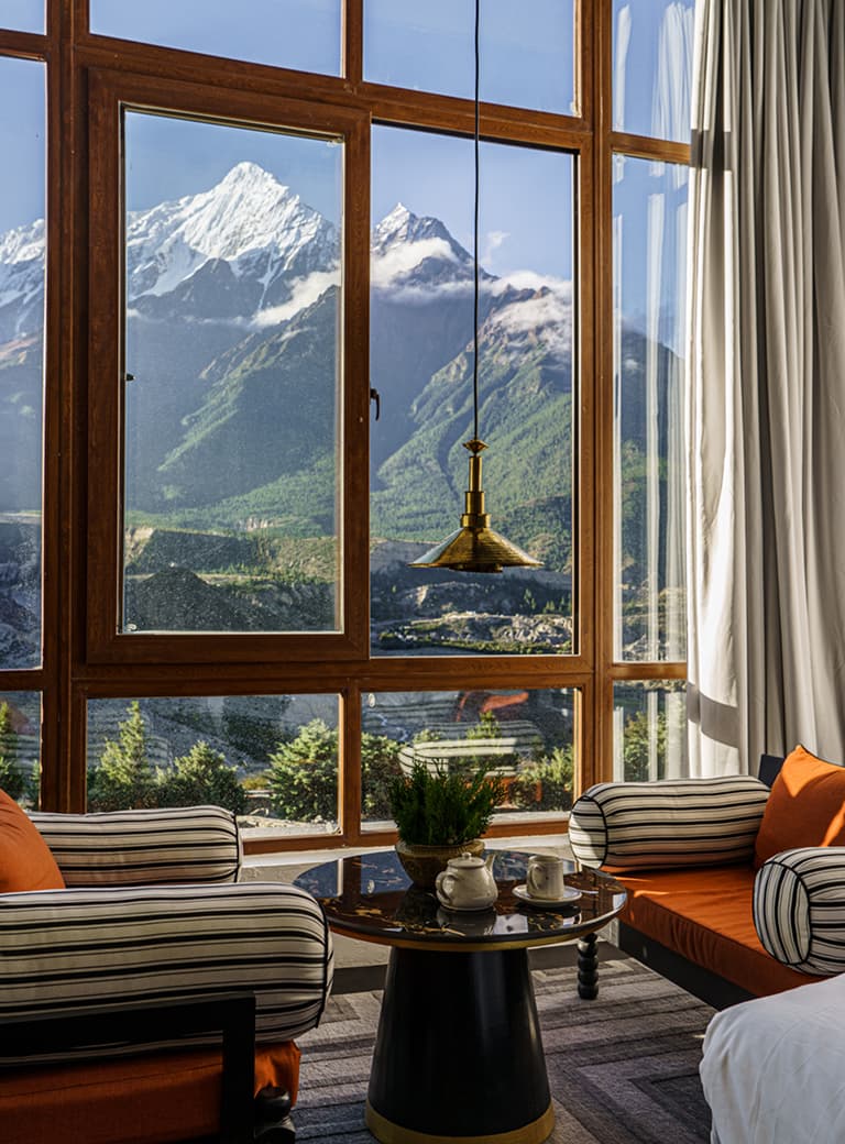 Cozy room with striped chairs, a small table, and a large window showcasing a majestic mountain landscape under a clear sky.