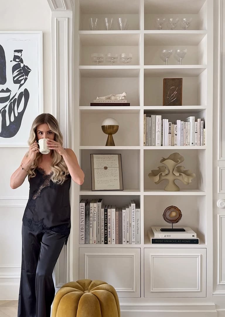 Woman in black outfit drinking coffee by a bookshelf with art, books, and decorative items in a stylish room.