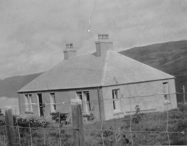 A vintage black-and-white photo of a small house with a hipped roof, chimneys, and a fence in a rural landscape with hills in the background.