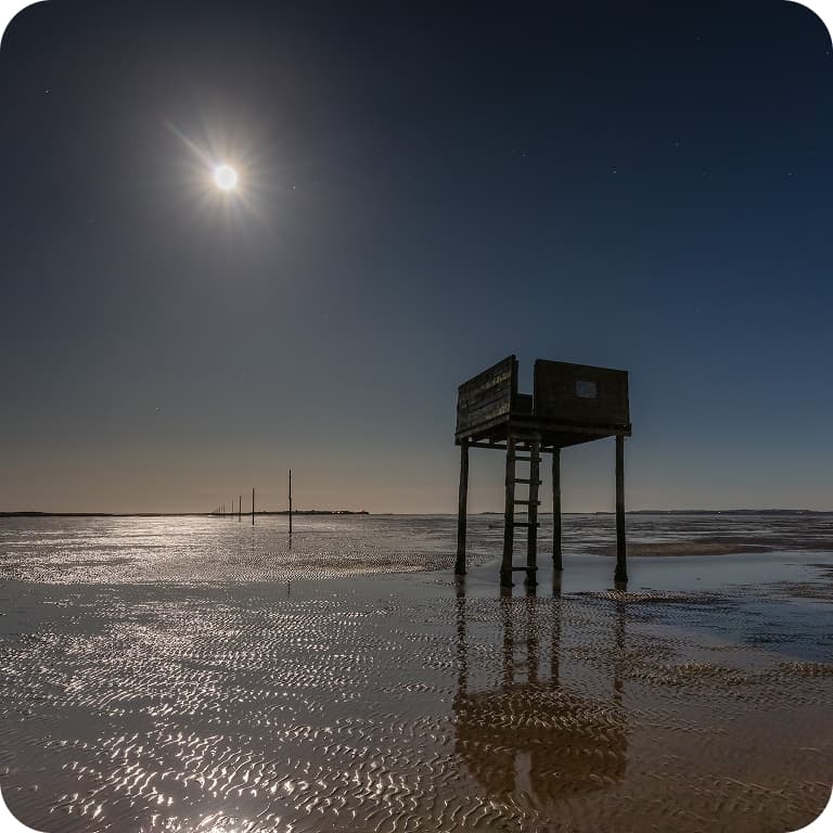 Moonlit beach scene with a wooden lifeguard tower on stilts, casting reflections on the wet sand.