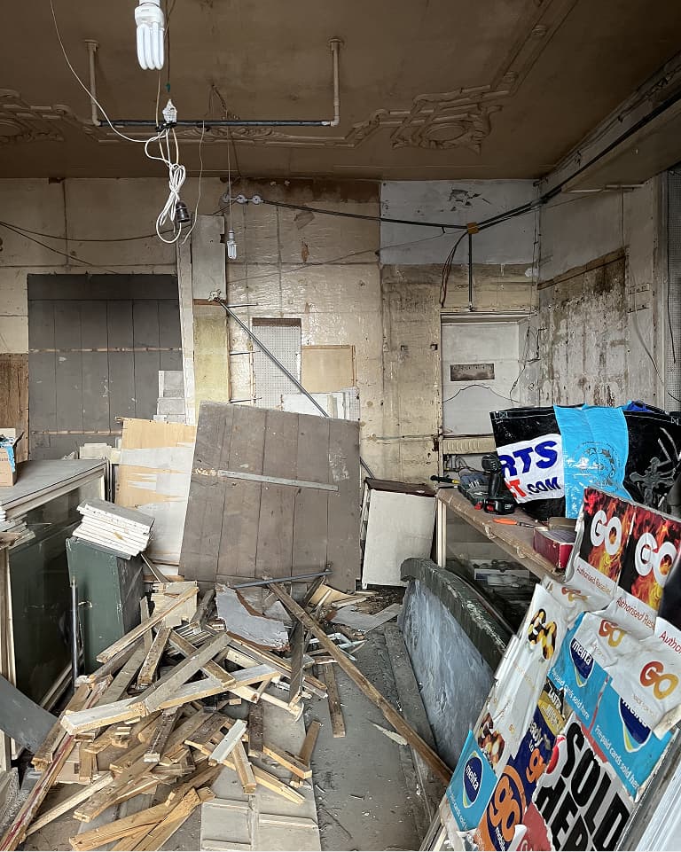 Cluttered interior with scattered wooden planks, old signs, and debris. Exposed wiring and peeling walls enhance the abandoned look.