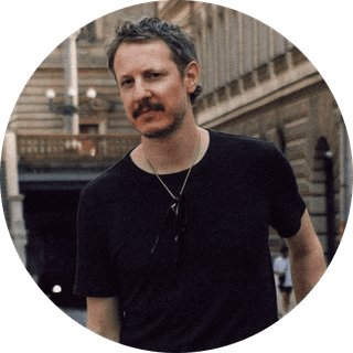 Man with a mustache wearing a black t-shirt and necklace, standing on a street with historic buildings in the background.