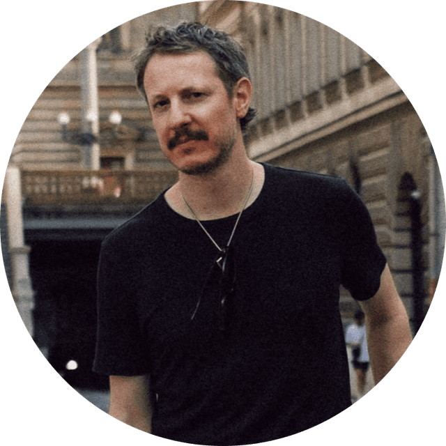Man with a mustache wearing a black t-shirt and necklace, standing on a street with historic buildings in the background.