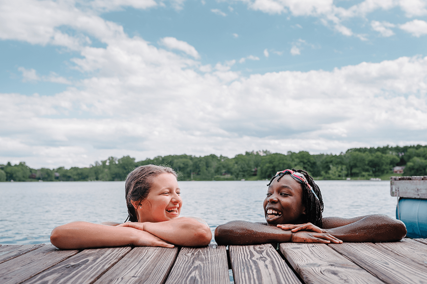Two young teenagers smiling and chatting while leaning on a wooden dock by a lake, with trees and a partly cloudy sky in the background.