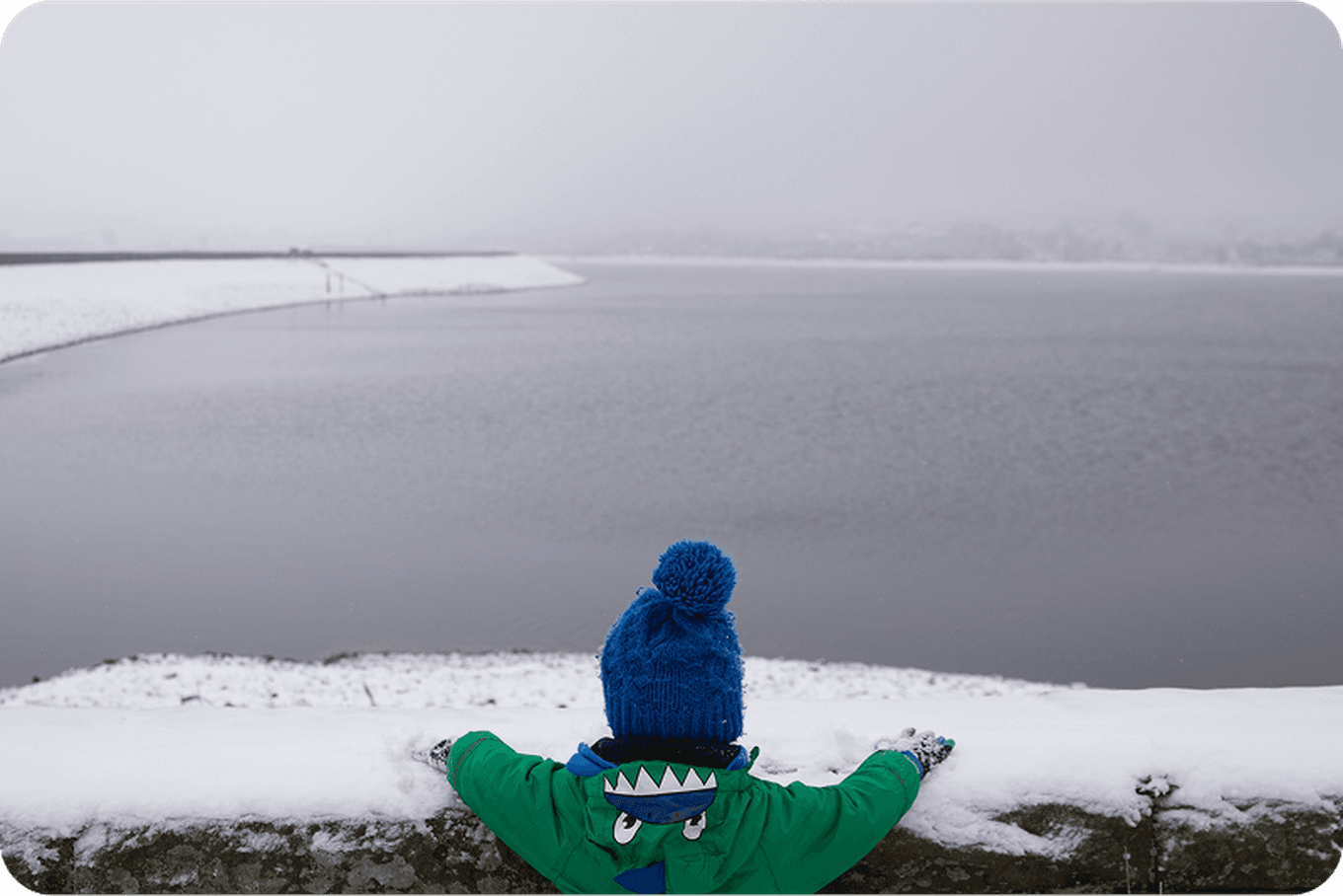 Child in a blue hat and green coat looks out over a snow-covered wall at a foggy, frozen lake.