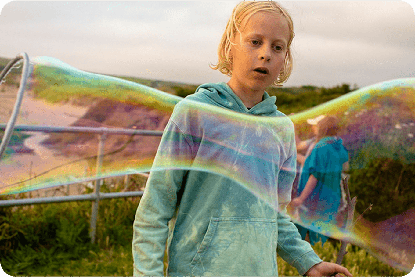 A child in a blue hoodie is mesmerized by a large, colorful soap bubble outdoors, with a blurred natural background.