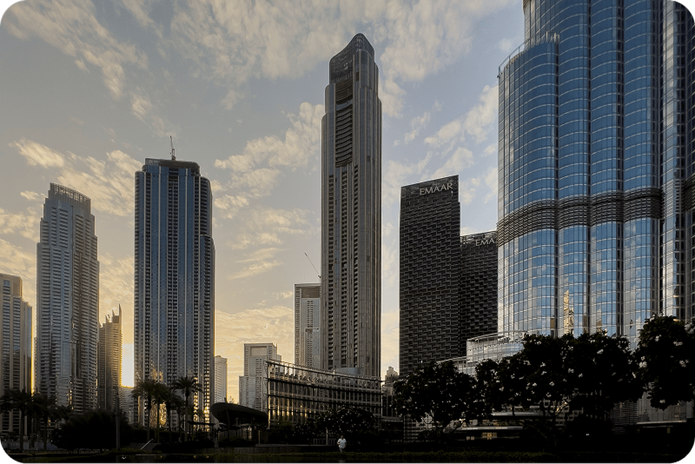 Skyline of modern skyscrapers with reflective glass facades against a sunset sky, surrounded by greenery and silhouetted trees.