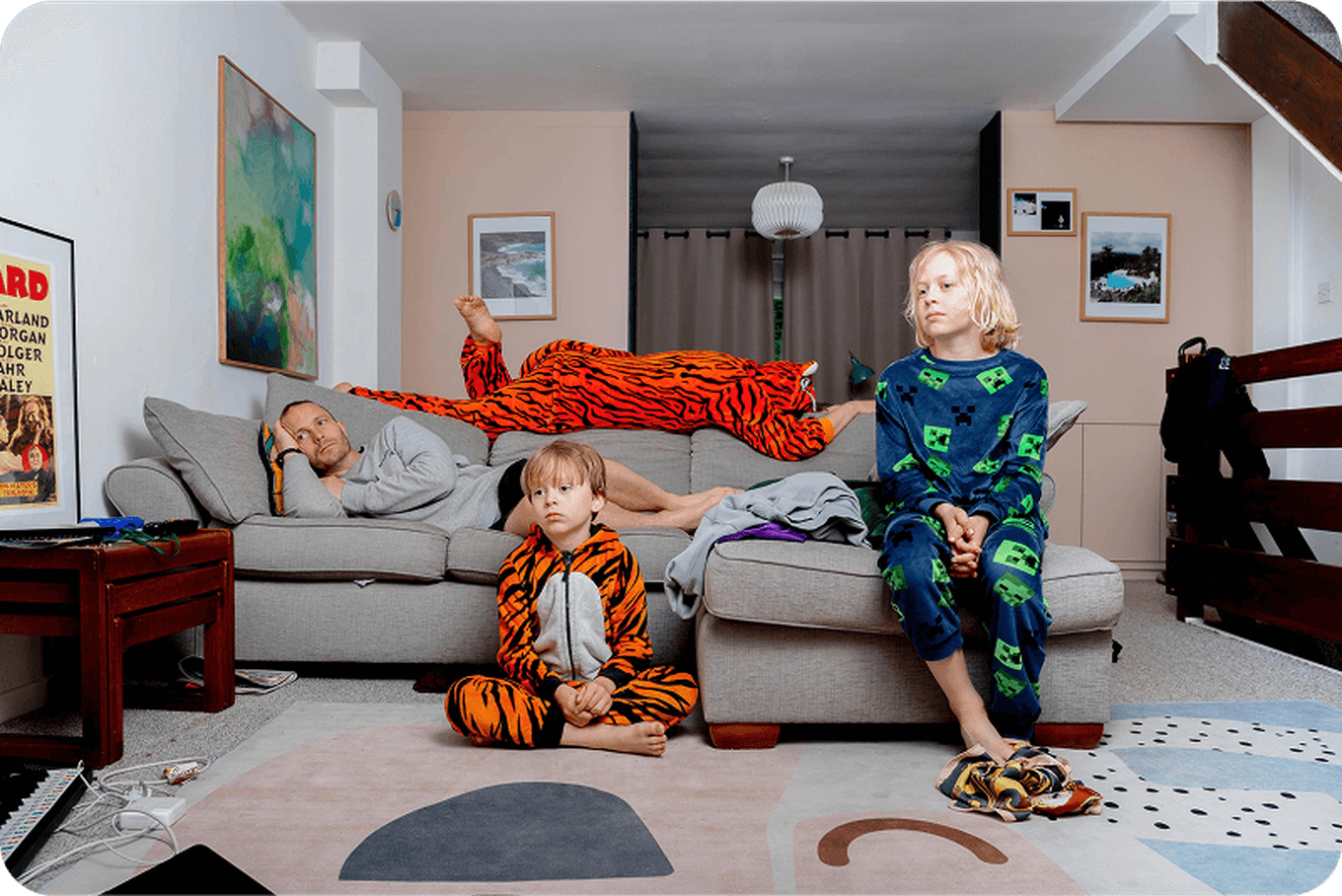 Three people in colorful pajamas relax in a living room. One lies on a couch, while two sit on the floor, all watching something off-screen.