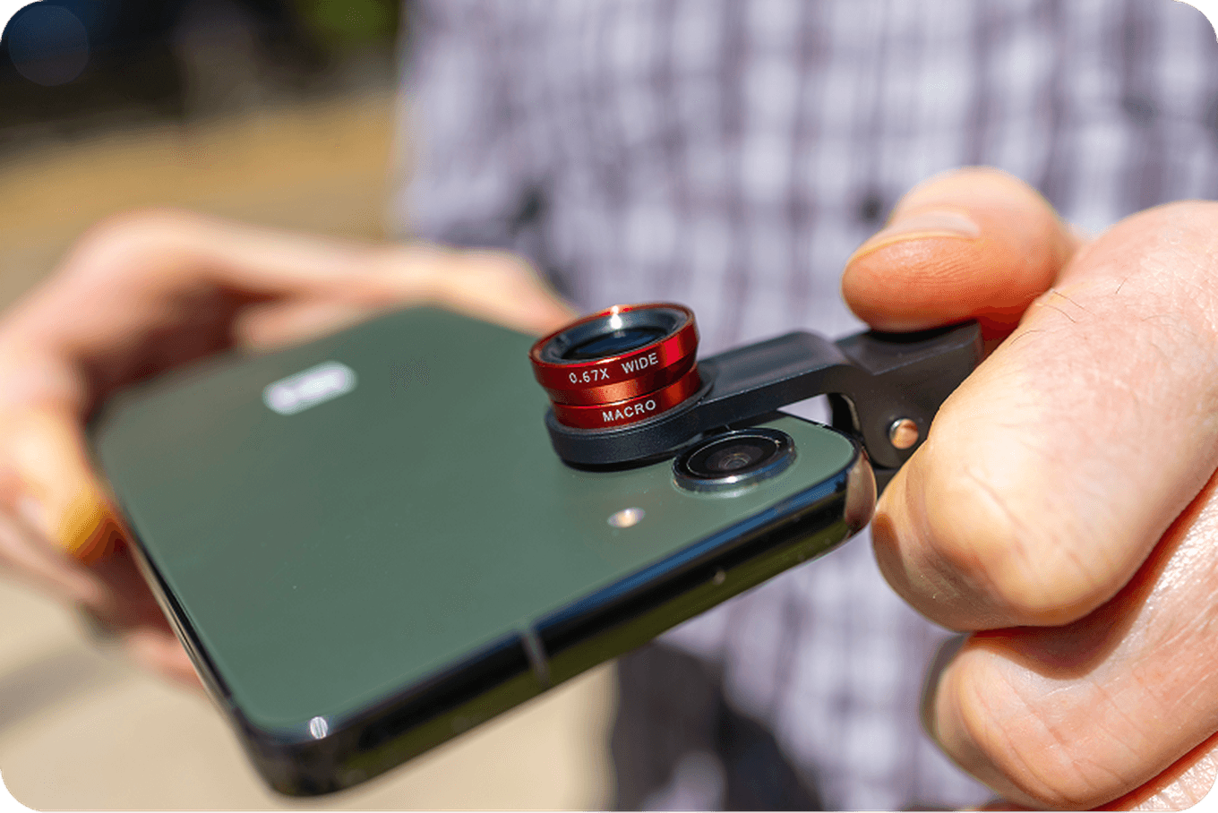 Close-up of hands attaching a red macro and wide-angle lens clip to a smartphone camera outdoors, with a blurred background.