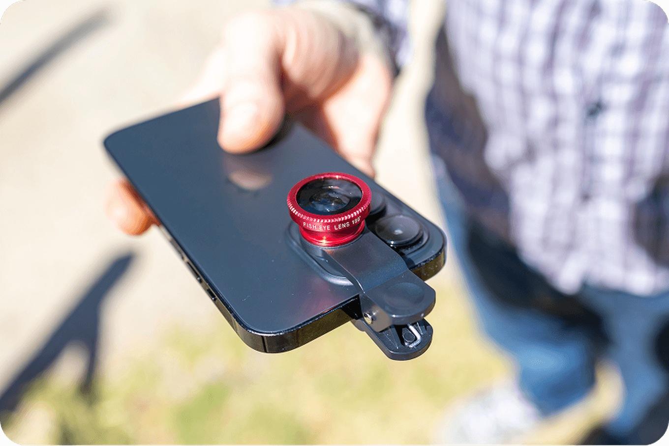 Person holding a smartphone with a red clip-on lens attached, outdoors in sunlight.