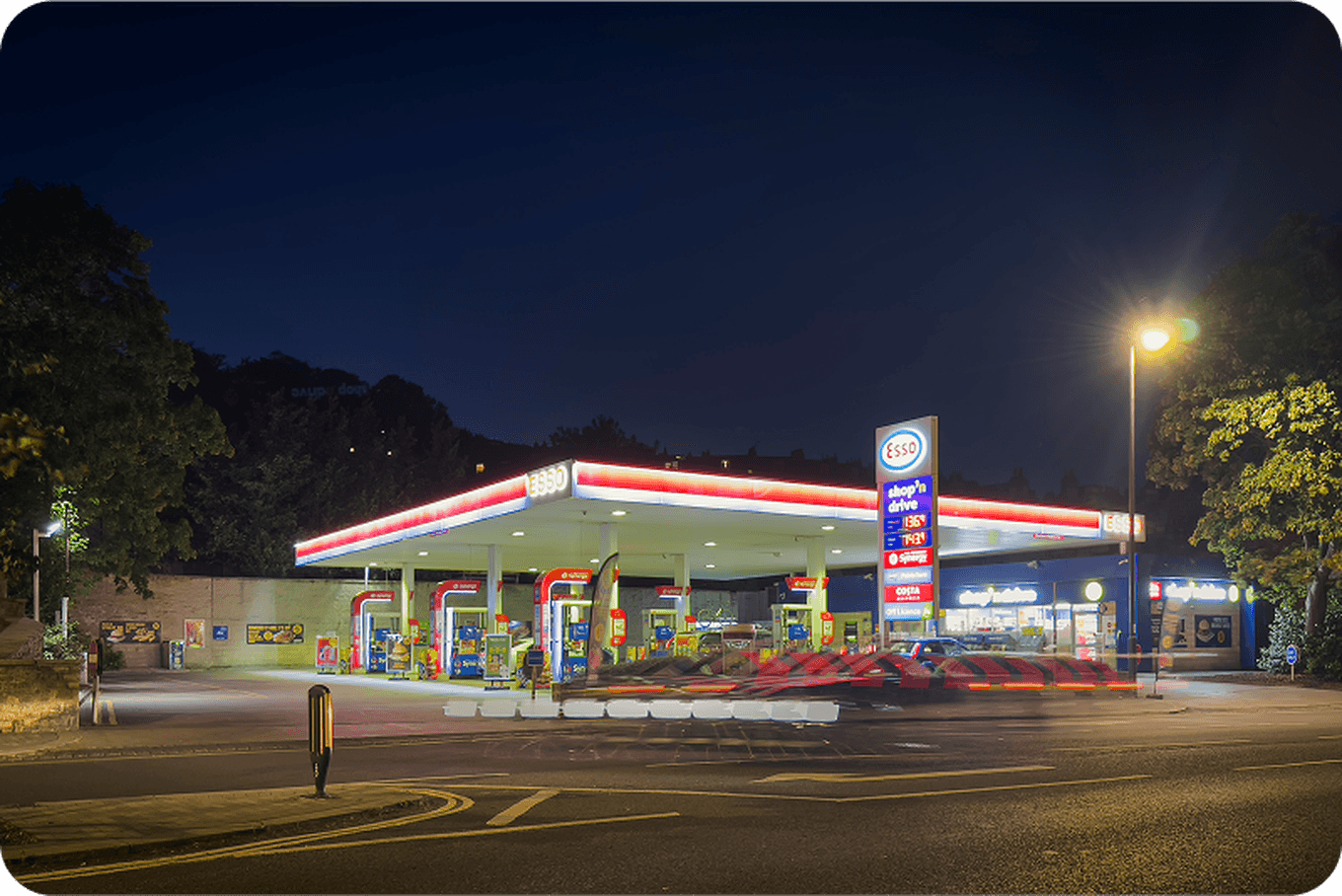 Night view of a brightly lit gas station with a convenience store, surrounded by trees and a road with light trails from passing vehicles.
