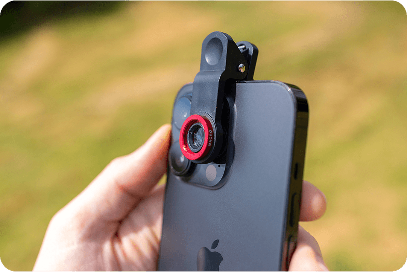 A hand holds a smartphone with an attached clip-on lens, featuring a red ring, enhancing the phone's camera capabilities in an outdoor setting.