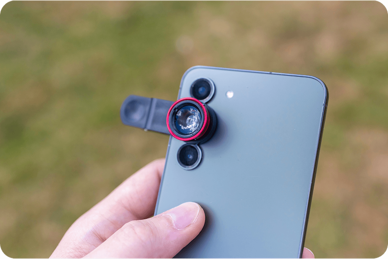 A person holds a smartphone with a red clip-on lens attached to the camera, set against a blurred outdoor background.