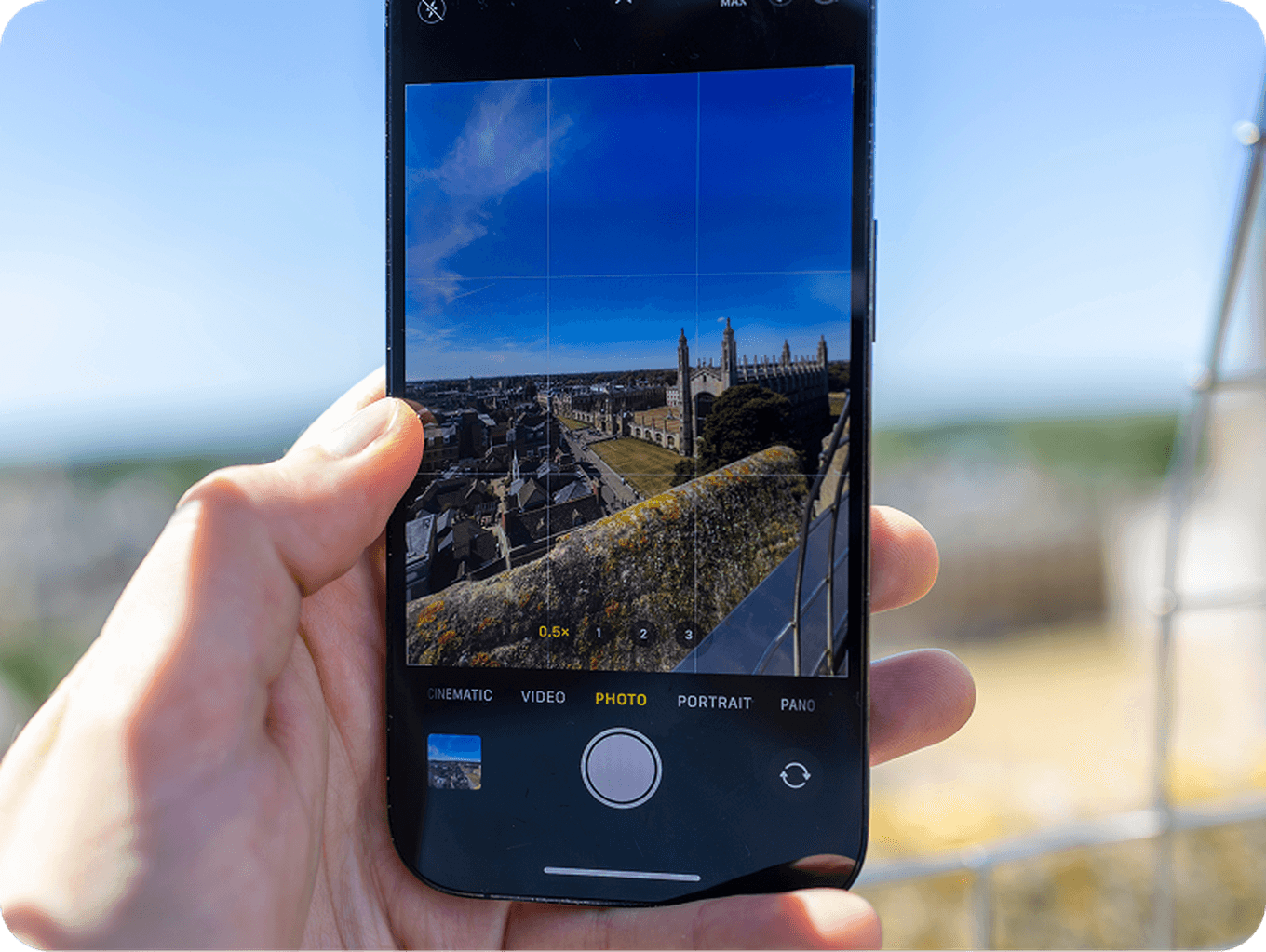 A hand holds a smartphone, capturing a scenic cityscape with historic buildings under a blue sky, displayed on the phone screen.