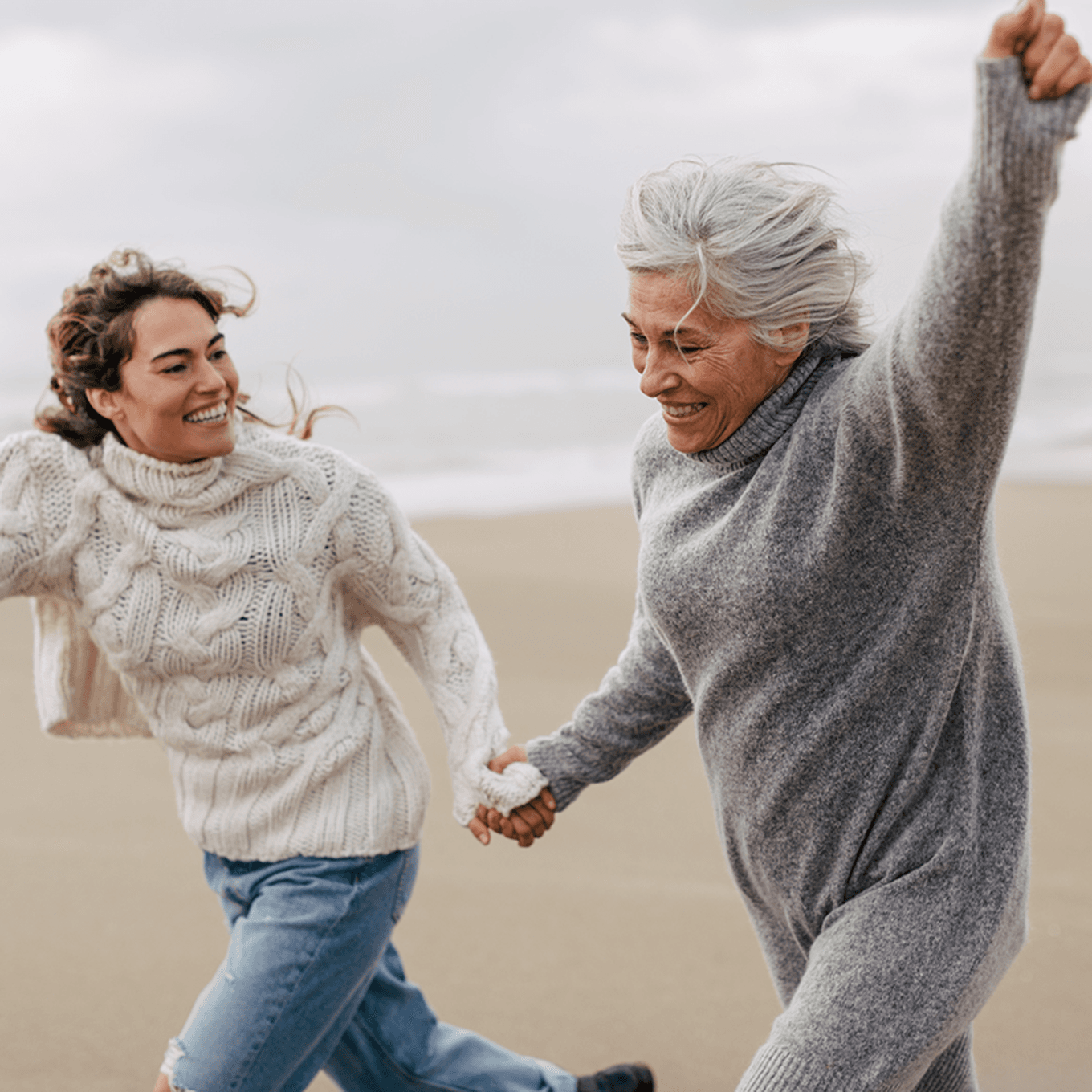 A senior mother and her adult daughter running on the beach holding hands