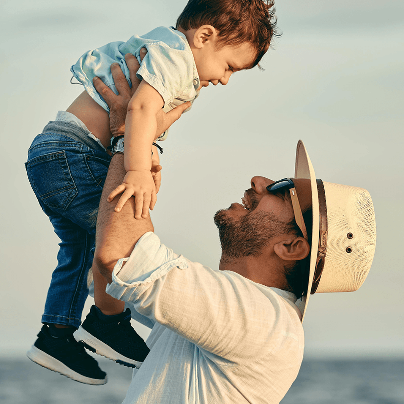 A man in a hat joyfully lifts a smiling child in the air by the seaside, both wearing casual clothing.