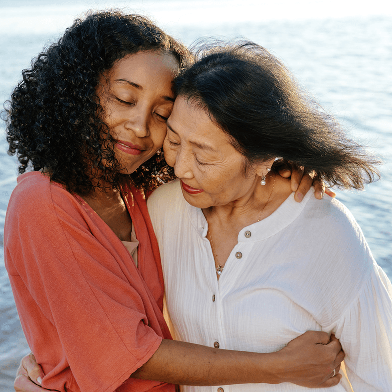 Two women embrace warmly by the seaside, eyes closed, exuding a sense of peace and affection.