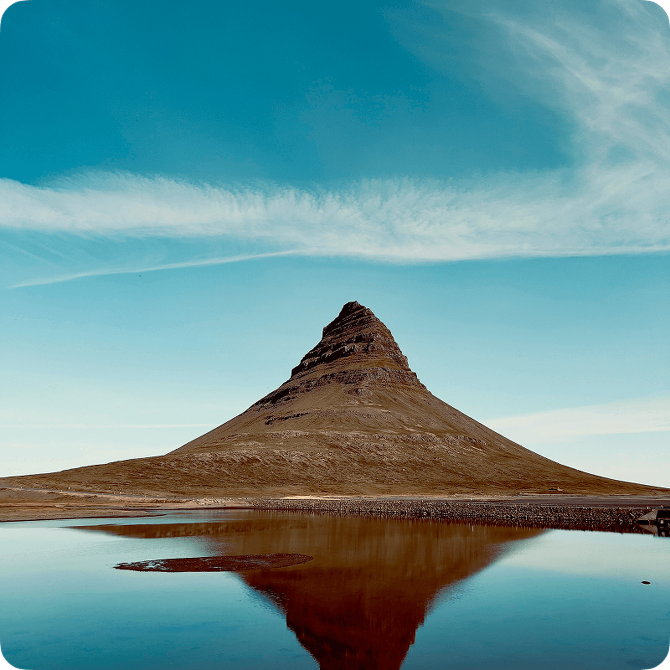 A conical mountain reflected in a calm body of water under a clear blue sky with wispy clouds.