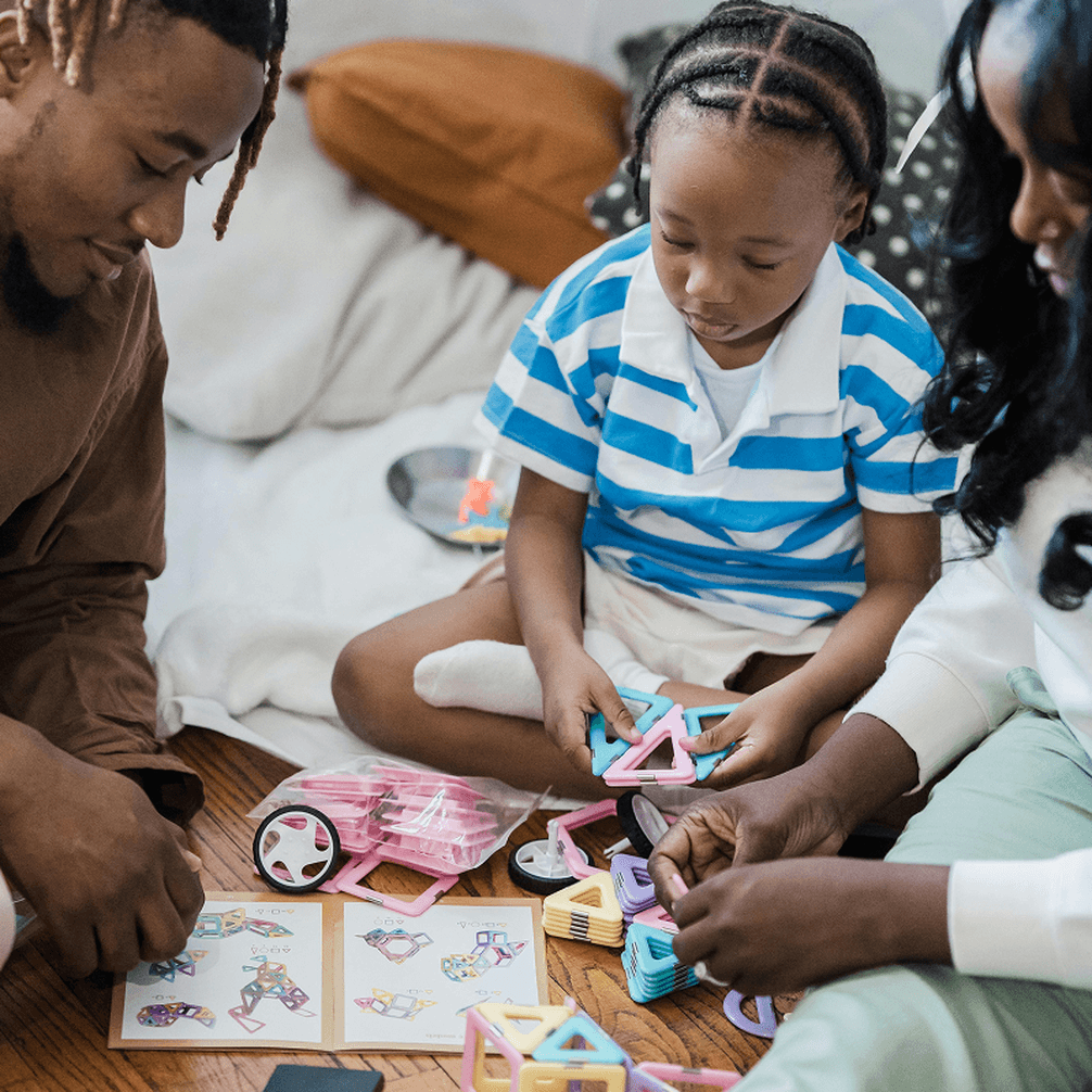 A family sits on the floor, playing with colorful magnetic building blocks. A child in a striped shirt focuses on assembling shapes.