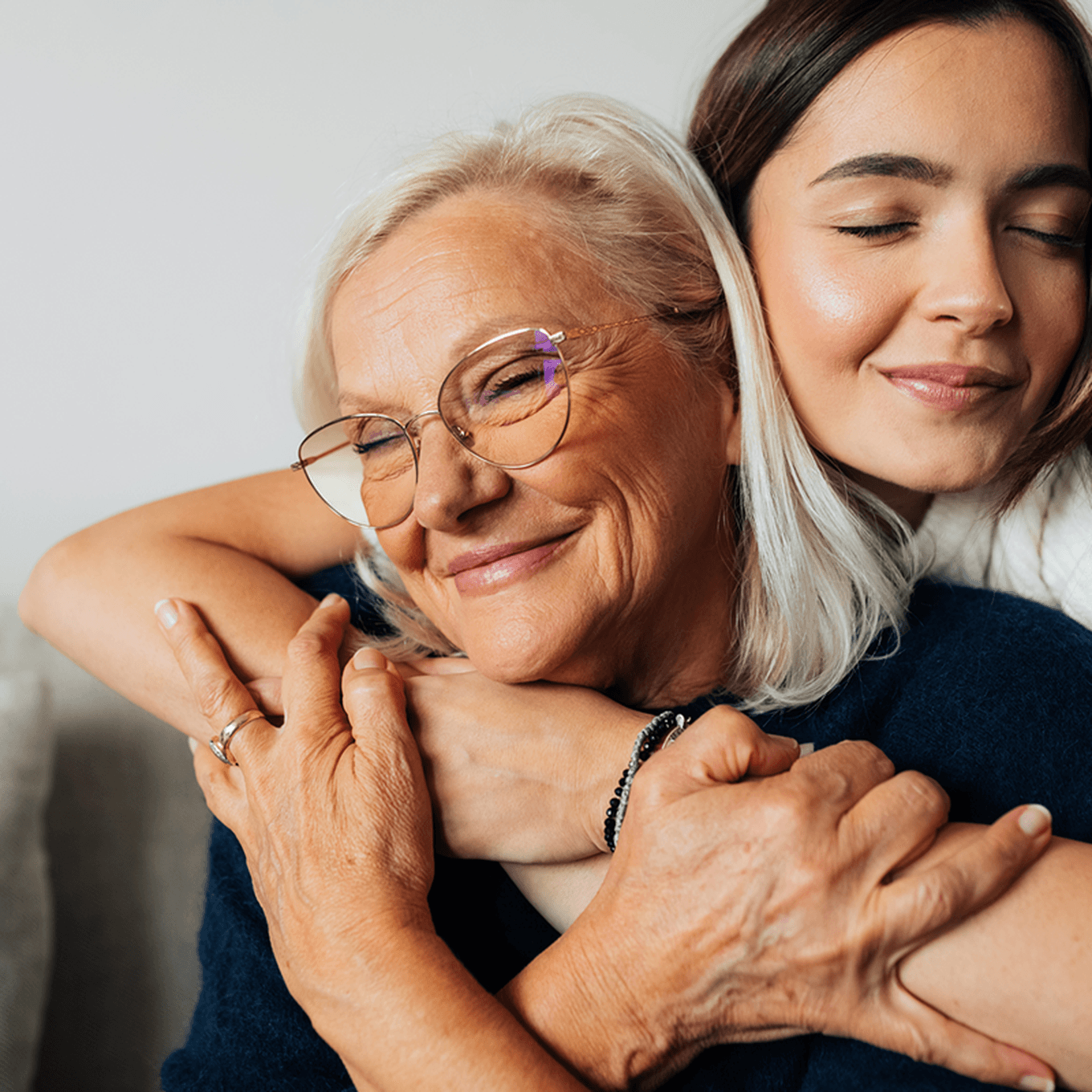 Elderly woman with glasses smiles warmly as a young woman hugs her from behind, both with eyes closed in a loving embrace.
