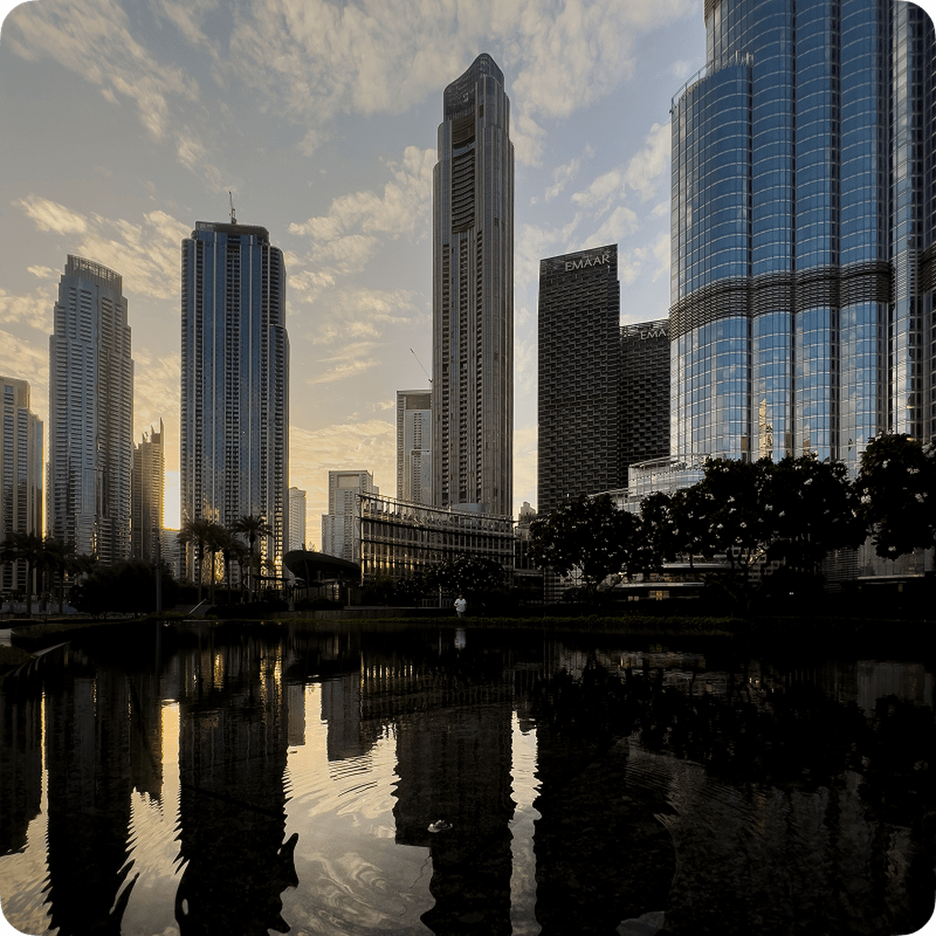 City skyline at sunset with tall skyscrapers reflecting in a calm water surface, surrounded by trees and a partly cloudy sky.