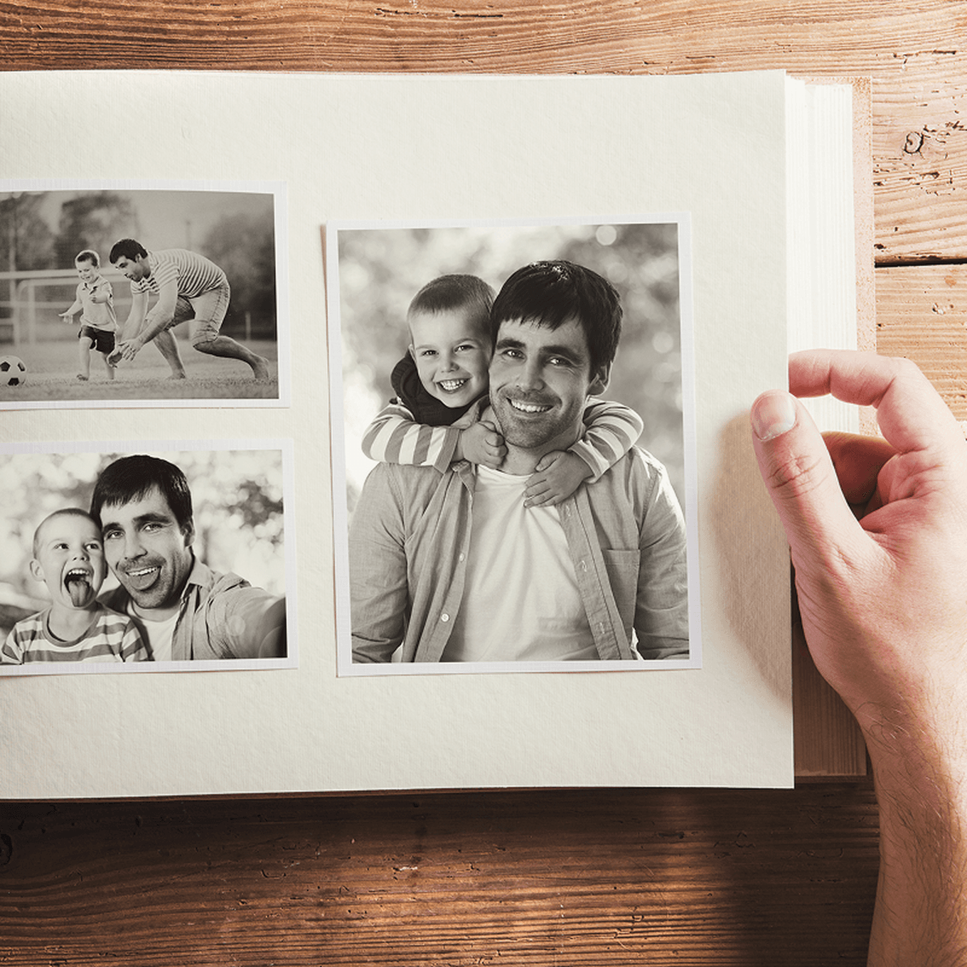 A hand holds an open photo album showing black-and-white photos of a smiling man and child in various playful moments.