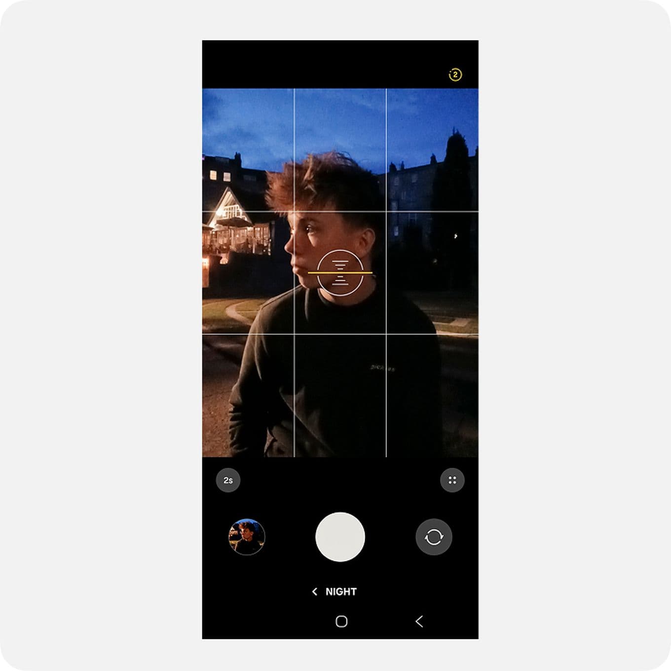 A smartphone screen displaying a camera app interface, capturing a person outdoors at night with buildings illuminated in the background.