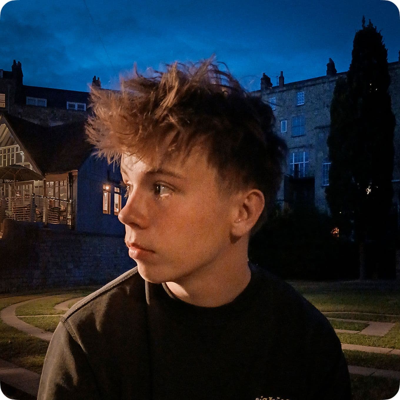 Teen boy with tousled hair gazes to the side, sitting outdoors at dusk. Dimly lit buildings and a deep blue sky form the background.