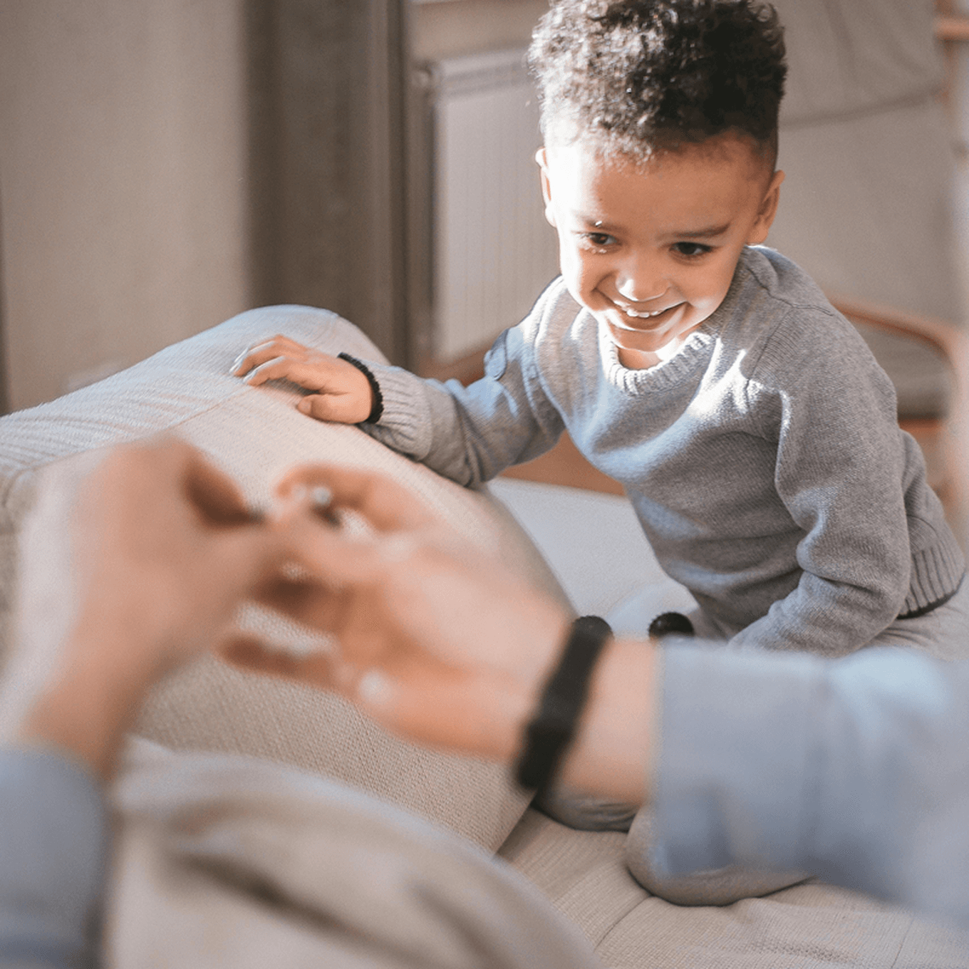 A smiling child in a grey sweater leans on a couch, looking at an adult's hand holding a small object in the foreground.