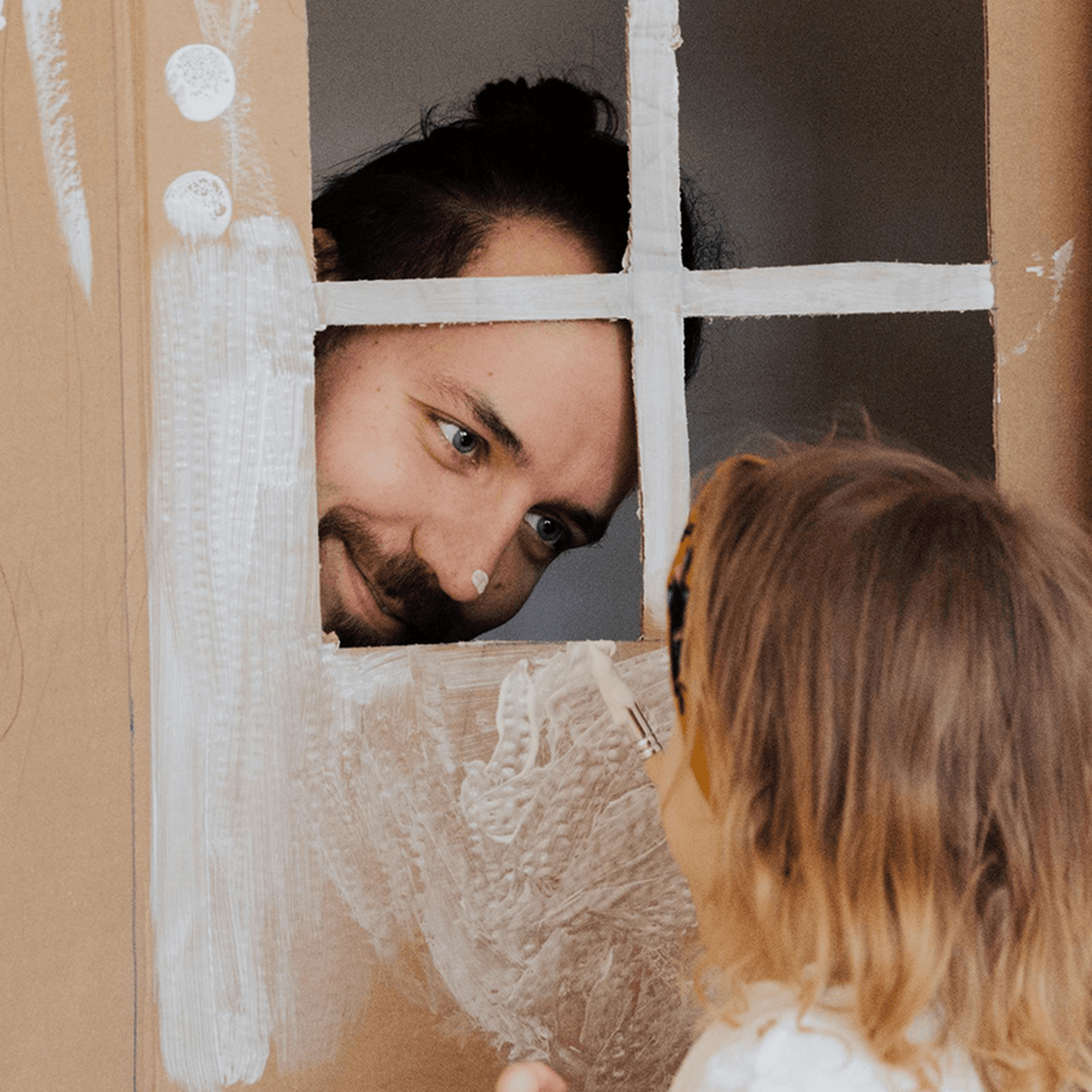 Man smiling through a cardboard window at a child painting the cardboard.
