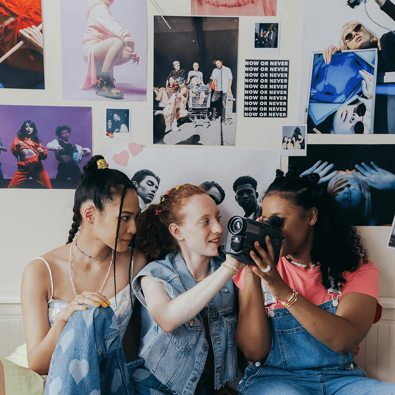 Three teenage friends sitting together, looking at an old-fashioned camcorder. The wall behind them is covered with 90s music posters