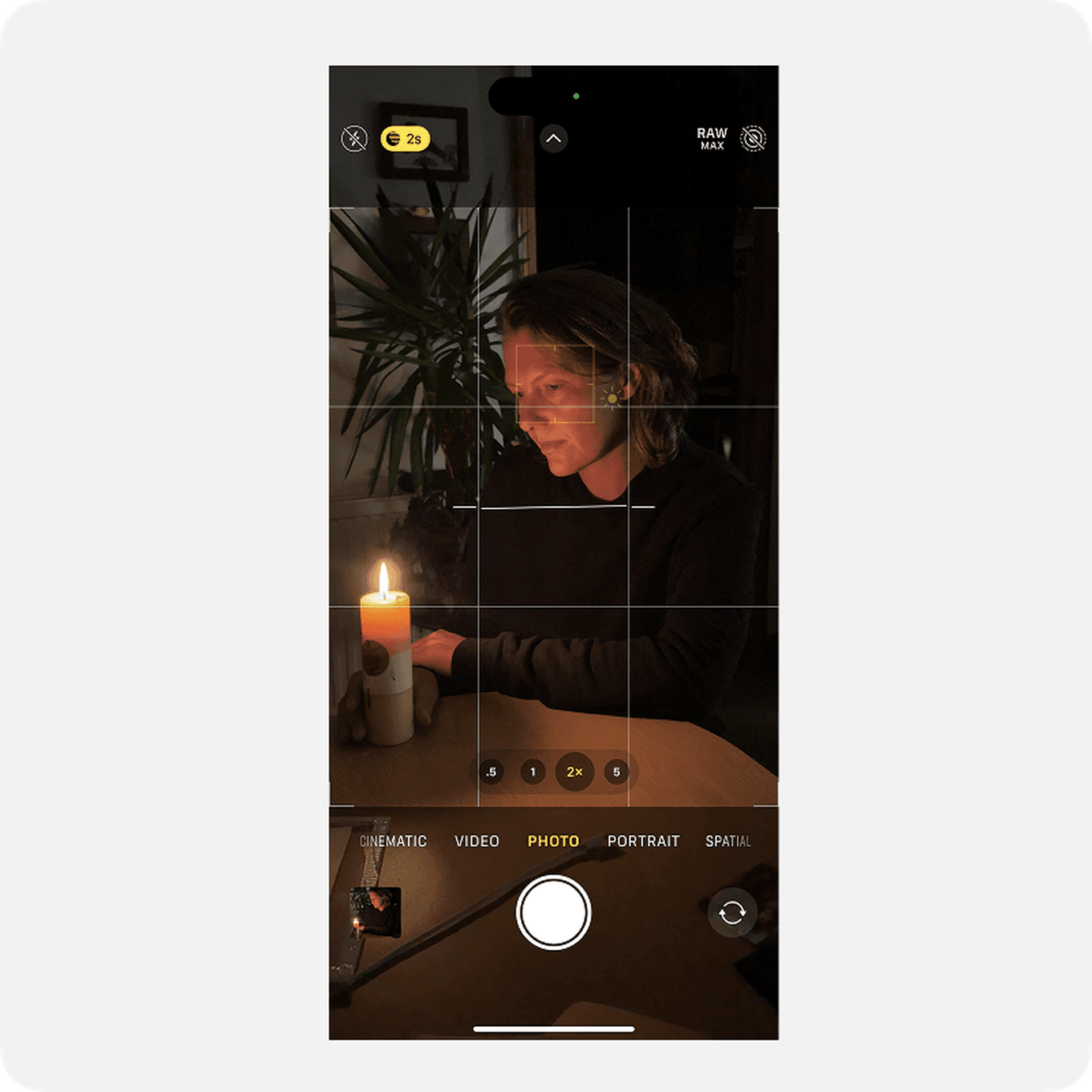 A person sits at a table in dim light, holding a lit candle. The scene is captured on a smartphone camera screen.