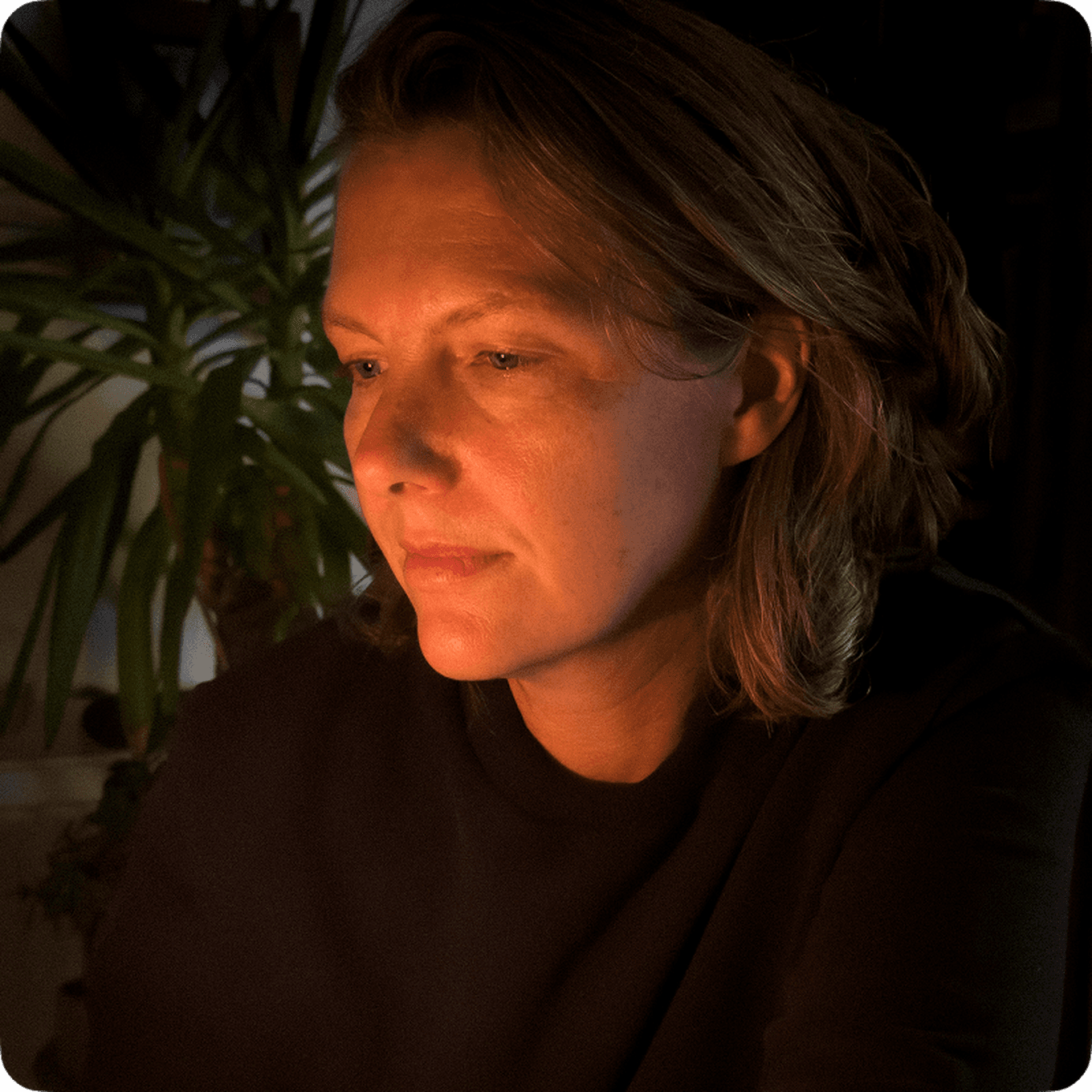 Person with short hair gazing thoughtfully, illuminated by soft light, with a large leafy plant in the background.