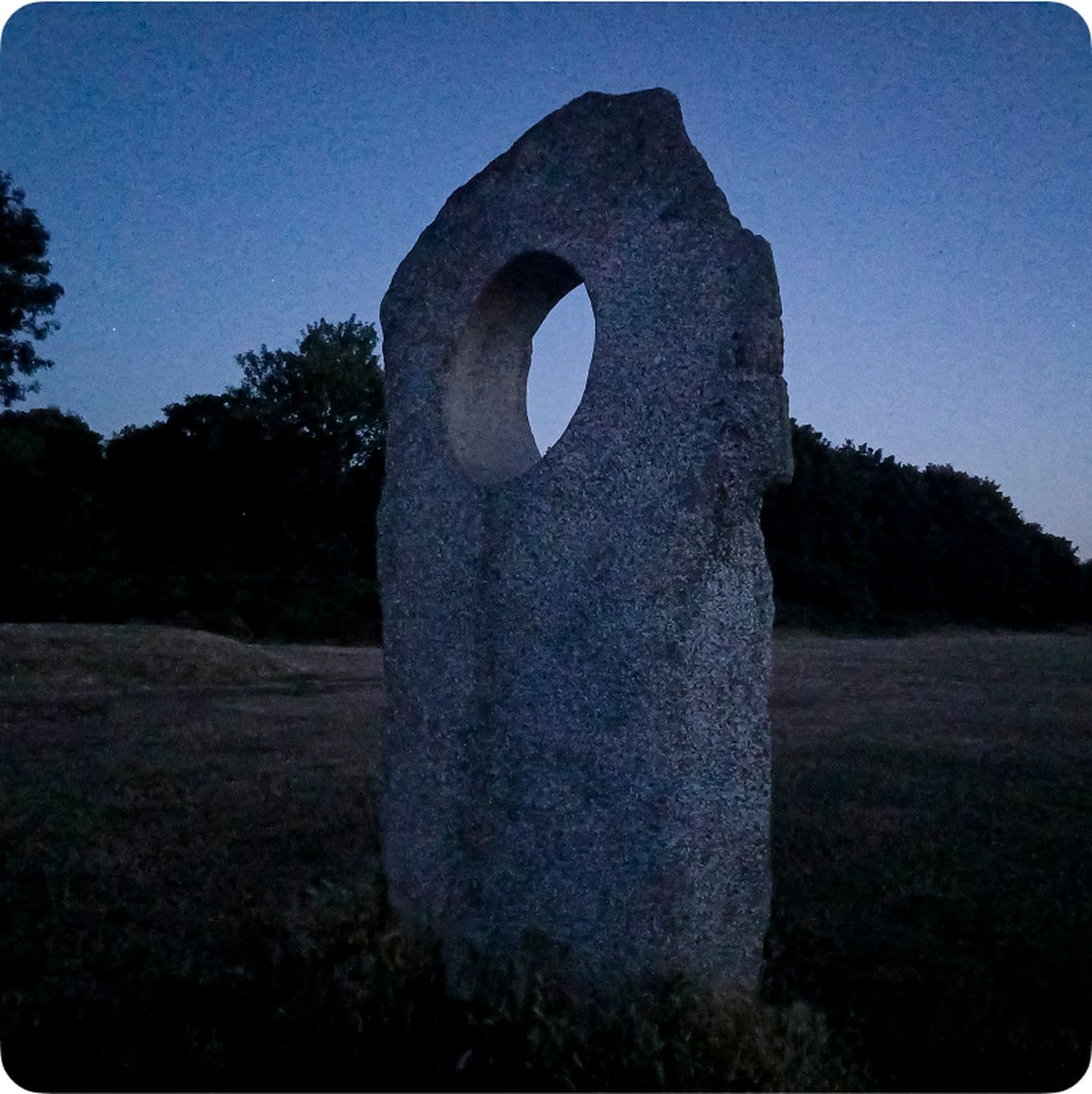 Stone monolith with a circular hole stands against a twilight sky, surrounded by grass and silhouetted trees.