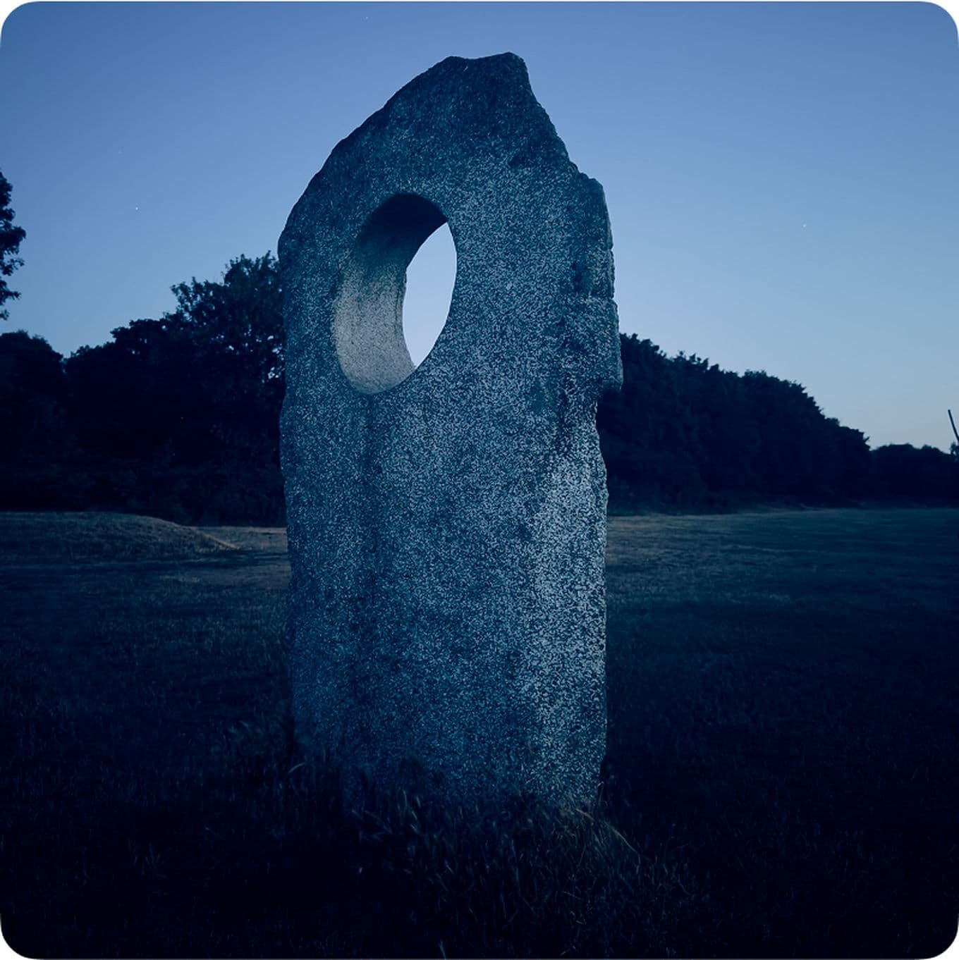 A tall, weathered stone slab with a circular hole stands in a grassy field at dusk, surrounded by trees.