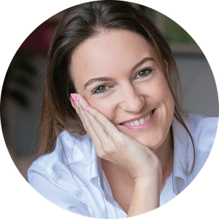 Smiling woman with brown hair resting her face on her hand, wearing a white shirt, close-up portrait.