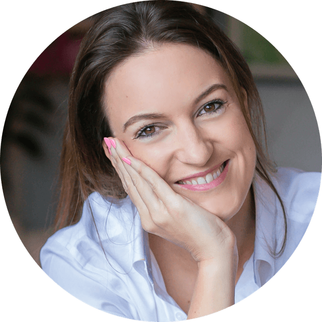 Smiling woman with brown hair resting her face on her hand, wearing a white shirt, close-up portrait.