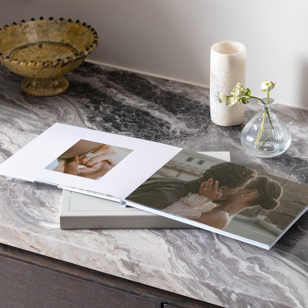An engagement photo book with layflat pages sits open on a marble counter, the photo book is open on a photo of a couple kissing and of the woman's engagement ring