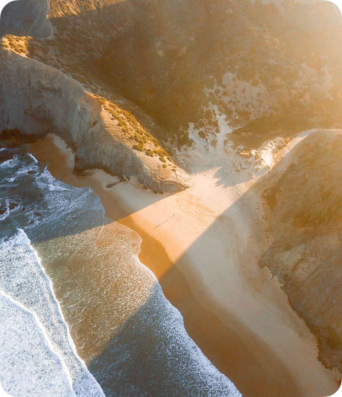 Aerial view of a secluded beach surrounded by rocky cliffs, with sunlight casting long shadows and waves gently lapping the shore.