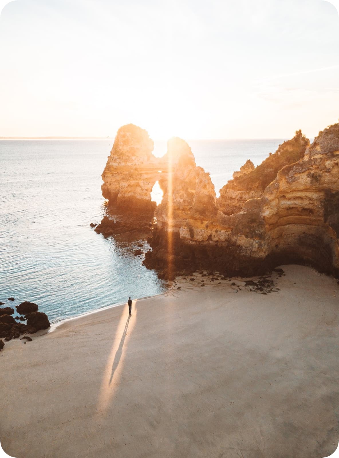 Sunrise over a rocky arch on a serene beach, casting long shadows on the sand with a lone person standing near the water's edge.
